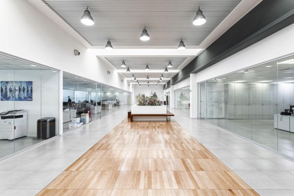 Vista del Corridoio Centrale. Una vista del corridoio centrale che separa gli uffici. Il pavimento sopraelevato garantisce una facile gestione degli impianti sottostanti. La combinazione di pavimentazione in piastrelle grigie e inserti in legno crea un effetto visivo piacevole e moderno.