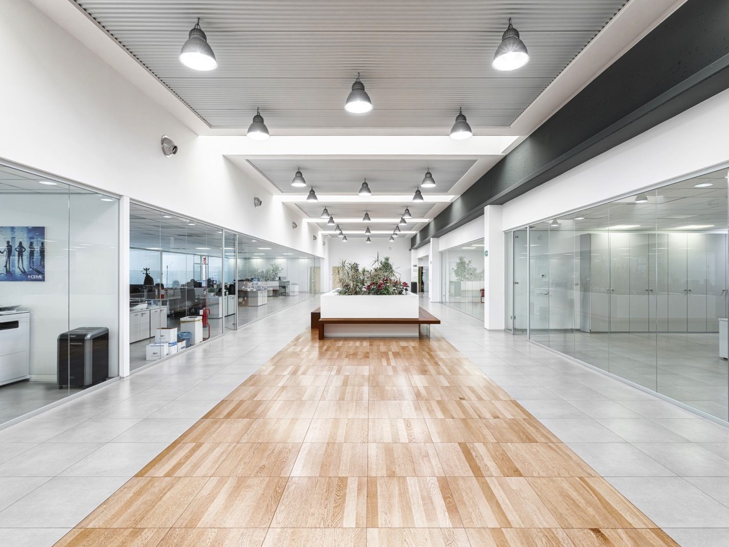 Vista del Corridoio Centrale. Una vista del corridoio centrale che separa gli uffici. Il pavimento sopraelevato garantisce una facile gestione degli impianti sottostanti. La combinazione di pavimentazione in piastrelle grigie e inserti in legno crea un effetto visivo piacevole e moderno.