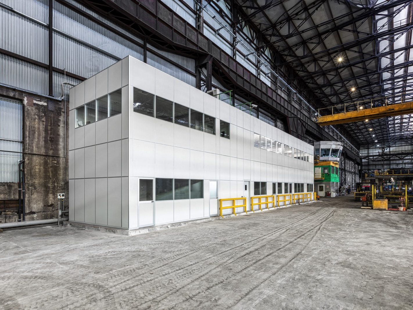 La vista esterna mostra una struttura moderna e funzionale, inserita all'interno di un contesto industriale. La facciata è composta da pannelli modulari di colore grigio, con finestre distribuite lungo tutto il perimetro superiore per garantire una buona illuminazione naturale all'interno. La struttura si distingue per la sua semplicità e linearità, contribuendo a un ambiente di lavoro efficiente e ben organizzato.