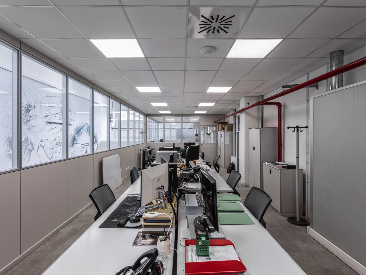 Vista interna di un ufficio al piano superiore Caratterizzato da ampie finestre che permettono la visibilità sull'area di produzione sottostante. Gli arredi sono funzionali e moderni, con scrivanie bianche e sedie ergonomiche.
