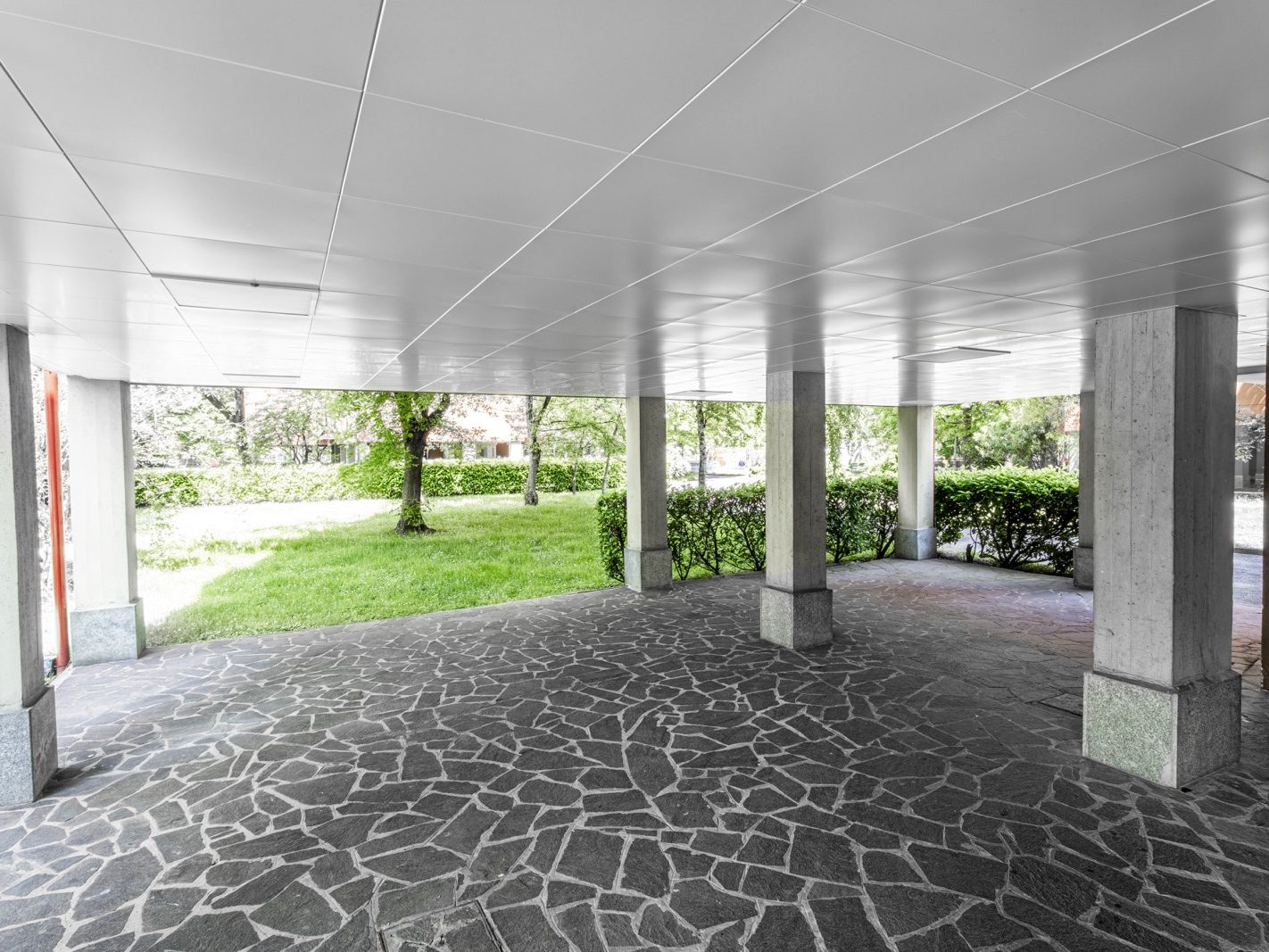 Un'altra vista dell'area di passaggio coperta che mette in evidenza la continuità del design e l'armonia tra soffitti e pavimenti. Si nota anche la vegetazione circostante, che contribuisce a creare un ambiente accogliente e rilassante.